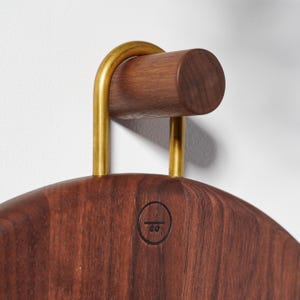 Pode incluir: Um cabide de madeira castanho escuro com uma pega de metal cor de latão. O cabide tem uma forma arredondada e um pequeno logótipo circular é visível na frente. A pega é curva e presa a um pino de madeira.