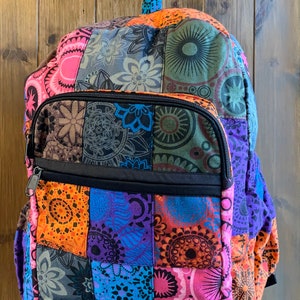 May include: A colorful patchwork backpack with a black zippered front pocket. The backpack features a variety of patterned squares in shades of pink, orange, blue, purple, and green. The bag has a top handle and adjustable straps.