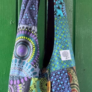 Op de afbeelding: Een hobo-tas met een patchworkontwerp met diverse cirkelvormige en geometrische patronen in tinten blauw, groen, paars en zwart. De tas heeft een lange schouderband en een klein wit vierkant label met een boomafbeelding en tekst.