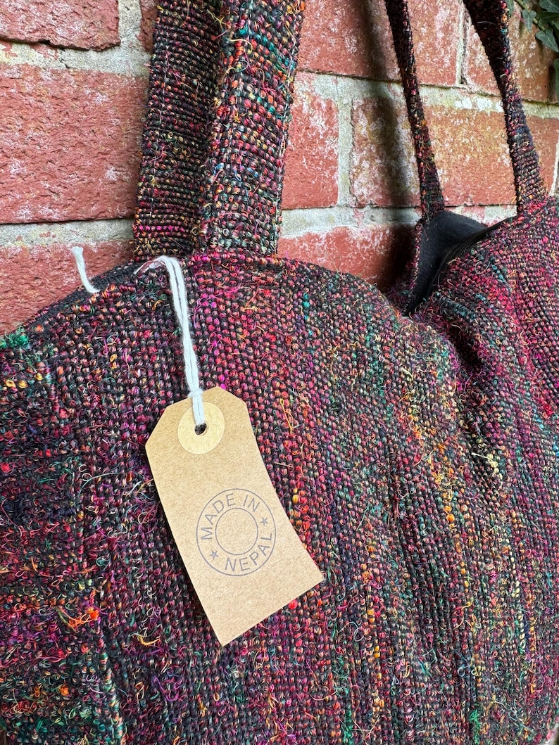 K&ouml;nnte beinhalten: Eine schwarze, mehrfarbige gewebte Tragetasche mit einem braunen Etikett, auf dem "Made in Nepal" steht.