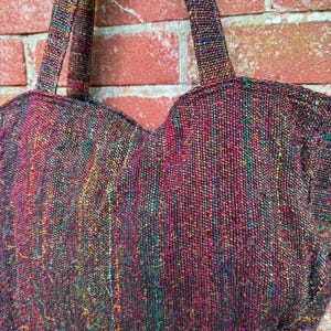 K&ouml;nnte beinhalten: Eine herzf&ouml;rmige Handtasche aus gewebtem Stoff mit einem mehrfarbigen Muster. Die Tasche hat zwei Henkel und ist aus einem dunklen Stoff mit roten, gr&uuml;nen und gelben Akzenten gefertigt.