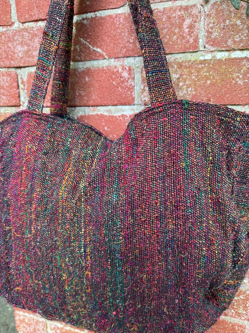 K&ouml;nnte beinhalten: Eine herzf&ouml;rmige Handtasche aus gewebtem Stoff mit einem mehrfarbigen Muster. Die Tasche hat zwei Henkel und ist aus einem dunklen Stoff mit roten, gr&uuml;nen und gelben Akzenten gefertigt.
