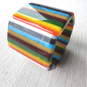 May include: A wide, rectangular cuff bracelet made of fused glass. The bracelet features horizontal, parallel stripes in vibrant colors including bright orange, yellow, deep green, dark brown, light blue, and white, with a smooth, glossy finish.