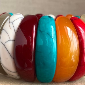 May include: A vibrant stretch bracelet with oval beads in various colours. The beads are red, orange, teal, white, and olive green. The bracelet has a diameter of approximately 5 cm.