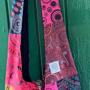 Puede incluir: Un bolso de hombro estilo hobo confeccionado con tela de patchwork que presenta una vibrante gama de colores, incluyendo naranja, rosa, morado y negro. El bolso muestra varios patrones circulares y florales, un cierre de botón de madera y una etiqueta.