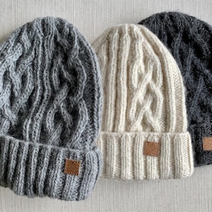May include: Three knitted cable knit hats in grey, white and black. Each hat has a leather tag with a logo.
