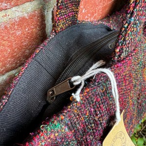 K&ouml;nnte beinhalten: Nahaufnahme einer bunten gewebten Tragetasche mit einer schwarzen Rei&szlig;verschlusstasche und einem braunen Etikett mit der Aufschrift "Made in Nepal".