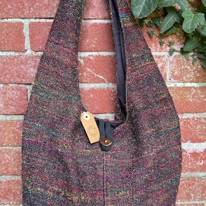 Op de afbeelding: Een zwarte geweven tote bag met een bruine leren knoopsluiting en een klein bruin label met de tekst "Made in Nepal". De tas heeft een veelkleurig geweven patroon met hints van rood, groen en blauw.