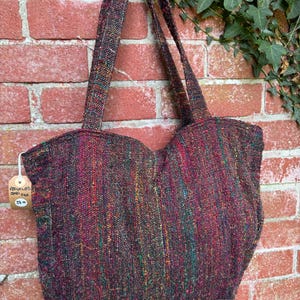 K&ouml;nnte beinhalten: Eine gro&szlig;e, farbenfrohe Tragetasche aus recycelter Sarisilk. Die Tasche hat eine gewebte Textur und ist in einem tiefen Burgunderrot mit gr&uuml;nlichen, goldenen und roten Akzenten gehalten. Die Tasche hat zwei lange Henkel und h&auml;ngt an einer Backsteinwand. Ein Preisschild ist an der Tasche befestigt, auf dem "Recycelte Sarisilk 25,00 &euro;" steht.