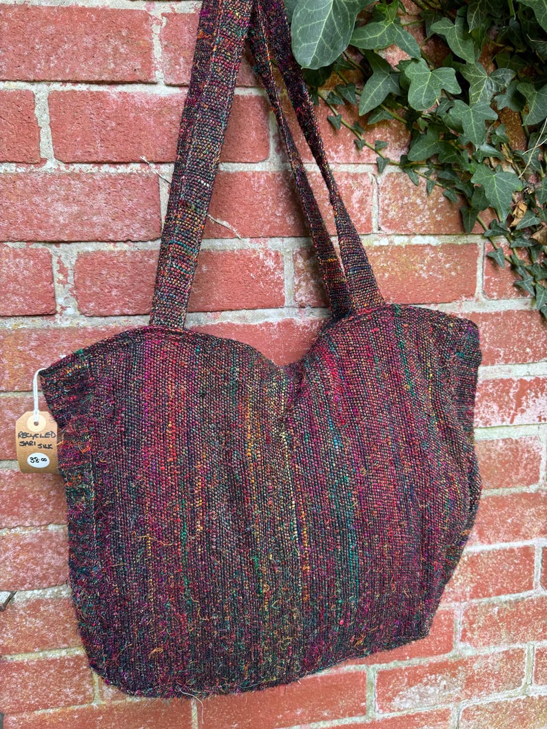 K&ouml;nnte beinhalten: Eine gro&szlig;e, farbenfrohe Tragetasche aus recycelter Sarisilk. Die Tasche hat eine gewebte Textur und ist in einem tiefen Burgunderrot mit gr&uuml;nlichen, goldenen und roten Akzenten gehalten. Die Tasche hat zwei lange Henkel und h&auml;ngt an einer Backsteinwand. Ein Preisschild ist an der Tasche befestigt, auf dem "Recycelte Sarisilk 25,00 &euro;" steht.