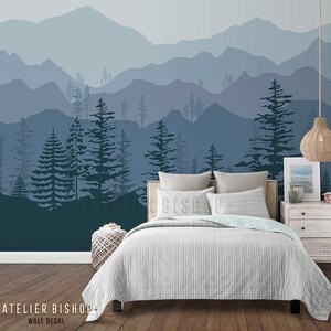 May include: A bedroom interior with a blue and grey mountain range wall decal featuring a silhouette of trees. The decal is above a bed with a white and grey striped duvet cover and a white headboard. A wicker pendant light hangs above a white bedside table with a framed print and a book.