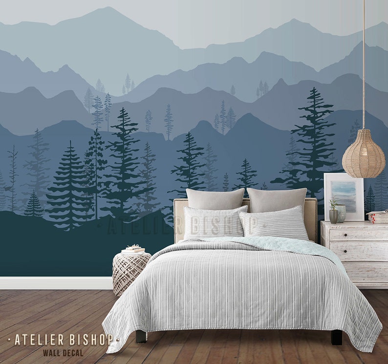 May include: A bedroom interior with a blue and grey mountain range wall decal featuring a silhouette of trees. The decal is above a bed with a white and grey striped duvet cover and a white headboard. A wicker pendant light hangs above a white bedside table with a framed print and a book.
