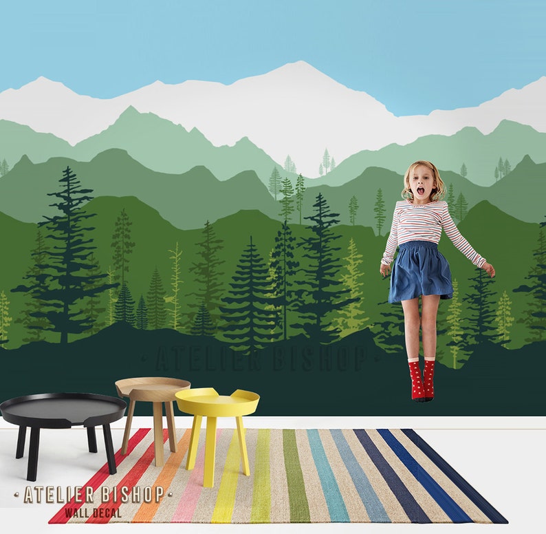May include: A green and blue mountain range wall decal with a silhouette of trees. A young girl is jumping in front of the decal. The decal is on a wall with a striped rug and a black and brown side table.