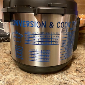 Op de afbeelding: Een Instant Pot van roestvrij staal met blauwe tekst die "Conversion & Cook Time" luidt en een tabel bevat voor het omrekenen van kooktijden van uren naar minuten. De tabel bevat kooktijden voor lage en hoge druk instellingen.