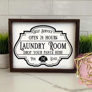 Op de afbeelding: Een ingelijst bord met zwarte tekst op een witte achtergrond met de tekst "Self Service Open 24 Hours Laundry Room Drop Your Pants Here Per 25c Load".