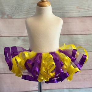 May include: A yellow and purple tulle tutu with multiple layers of ribbon. The tutu is on a white dress form.