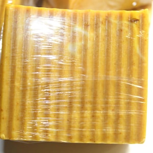 May include: A rectangular bar of soap wrapped in clear plastic. The soap is a golden-yellow colour with vertical ridges. The soap appears to be handmade and natural. The soap is a beauty product.