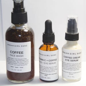 May include: Three glass bottles with black pump tops and labels. The bottles contain coffee face wash, vitamin C and coffee face and eye serum, and coffee cream eye serum. The labels are white with black text and the brand name "Urban Girl Sudz".