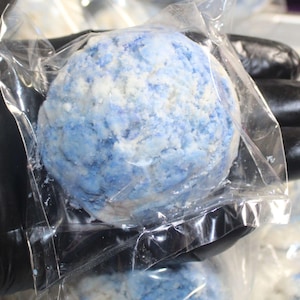 May include: A round, blue and white bath bomb is encased in clear plastic. The bath bomb has a marbled appearance with shades of blue and white. The bath bomb is held by a gloved hand.