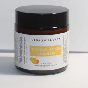 Könnte beinhalten: Ein braunes Glasgefäß mit schwarzem Deckel, das eine Gesichtsmaske enthält. Das weiße Etikett trägt den Text URBAN GIRL SUDZ, PUMPKIN + KAOLIN FACE MASK und 4 oz. / 120 g. Es enthält einen gelben rechteckigen Akzent und zwei kleine Kürbisse.