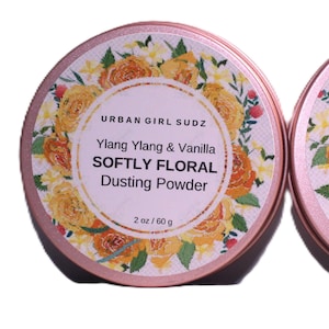 May include: A rose-gold tin of "SOFTLY FLORAL Dusting Powder" by URBAN GIRL SUDZ. The label features text and floral designs with the scent "Ylang Ylang & Vanilla". The container holds 60 g of powder.