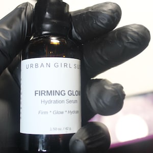 May include: A brown glass bottle of Urban Girl Suds Firming Glow Hydration Serum. The bottle has a black dropper and a white label with product information. The bottle is held by a gloved hand. The label states the serum contains 1.50 oz / 42 g.