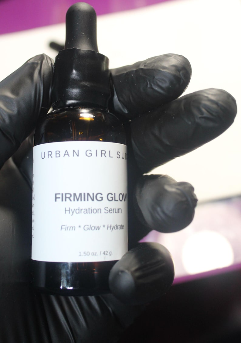 May include: A brown glass bottle of Urban Girl Suds Firming Glow Hydration Serum. The bottle has a black dropper and a white label with product information. The bottle is held by a gloved hand. The label states the serum contains 1.50 oz / 42 g.