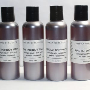 Może przedstawiać: Cztery butelki Pine Tar Body Wash firmy Urban Girl Sudz. Butelki mają metaliczny kolor malwy z czarnymi zakrętkami. Każda butelka ma białą etykietę z informacjami o produkcie, w tym składnikami takimi jak kwas salicylowy, aloes, drzewo herbaciane i cedr.