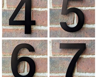 Peut inclure: Quatre numéros de maison en métal noir, 4, 5, 6 et 7, sont fixés à un mur en briques.