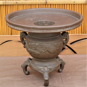 May include: A dark brown bronze vase with a wide, round top and three legs. The vase has a decorative design of stylized flowers and leaves.
