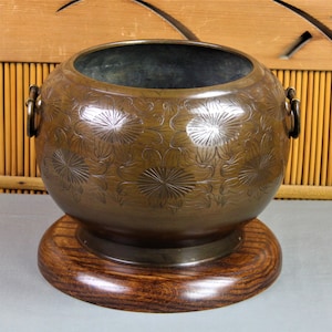 May include: A large, round, bronze planter with two handles and a floral design. The planter is sitting on a wooden base.