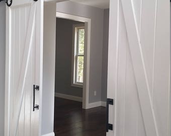 May include: Two white barn doors with black hardware and a black track. The doors are open and reveal a room with hardwood floors and a window.