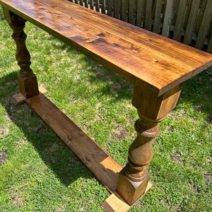 May include: A long, wooden console table with a dark brown stain. The table has a simple, rectangular top and turned legs with a square base. The table is sitting on a green lawn.