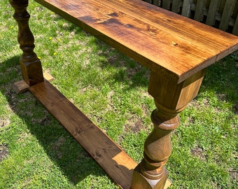 May include: A long, wooden console table with a dark brown stain. The table has a simple, rectangular top and turned legs with a square base. The table is sitting on a green lawn.