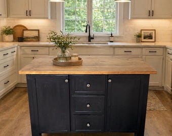 Puede incluir: Una isla de cocina con una encimera rectangular de madera marrón claro y una base negra con dos armarios laterales y tres cajones centrales. La isla está situada en una cocina con armarios blancos y una ventana sobre el fregadero.