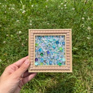 May include: A square wooden frame with a beaded border containing a dense arrangement of small, reflective sequins and beads in various shades of blue, green, and silver, creating a textured, shimmering mosaic effect within the frame.