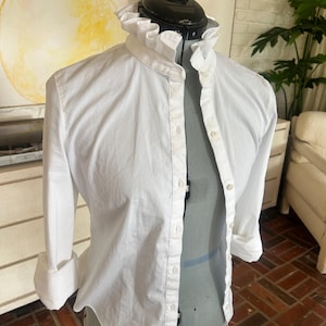 May include: A white button-down shirt with a ruffled collar and long sleeves, displayed on a gray dress form. The sleeves are cuffed at the elbows. The shirt is unbuttoned, revealing the form.