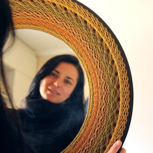 Op de afbeelding: Een ronde spiegel met een kleurrijk string art-ontwerp in tinten oranje, geel en groen. De spiegel reflecteert een vrouw met donker haar.