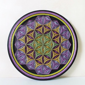 May include: A round, black wall art piece featuring a geometric design. The artwork is composed of intricate string art in shades of purple, yellow, and green, creating a symmetrical pattern. The piece has a black border with a purple and yellow accent.