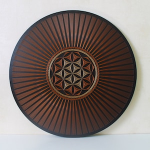May include: A round wooden wall art piece with a brown and gold geometric design in the center. The design is surrounded by a black background with brown lines radiating outward.