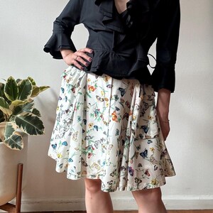 May include: A woman wearing a black ruffled blouse with a gold chain necklace and a white skirt with a butterfly print.