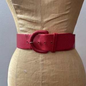 Puede incluir: Un cinturón ancho y rojo con una superficie texturizada y una hebilla grande y redondeada. El cinturón se muestra en un maniquí vintage. El color y el diseño llamativos del cinturón lo convierten en un accesorio llamativo.
