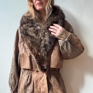 May include: A brown leather jacket featuring a fur collar. The jacket has a cinched waist and button details. The sleeves are long and the leather has a distressed look. The jacket is worn with blue jeans.