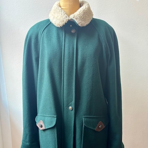May include: A dark green wool-blend coat featuring a cream-colored faux shearling collar, a front button placket with visible metal buttons, and two large patch pockets on the front, each accented with a brown leather-like triangular detail.
