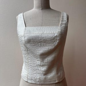 May include: Ivory-colored square-neck top with a textured, embossed pattern. The sleeveless top features a fitted silhouette and wide shoulder straps. The fabric has a subtle sheen, and the garment is displayed on a mannequin.