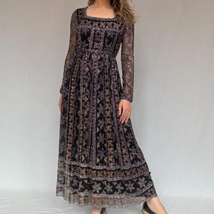 May include: A long black dress with a square neckline and long sleeves. The dress features a detailed pattern in gold and purple. The model is wearing black and gold shoes. The dress is a maxi length.