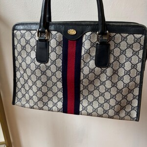 May include: A vintage handbag with a classic design. The bag features a repeating pattern of interlocking "G" logos in a beige and navy blue color scheme. A central stripe in red and navy blue adds a pop of color. The bag has black handles and trim.