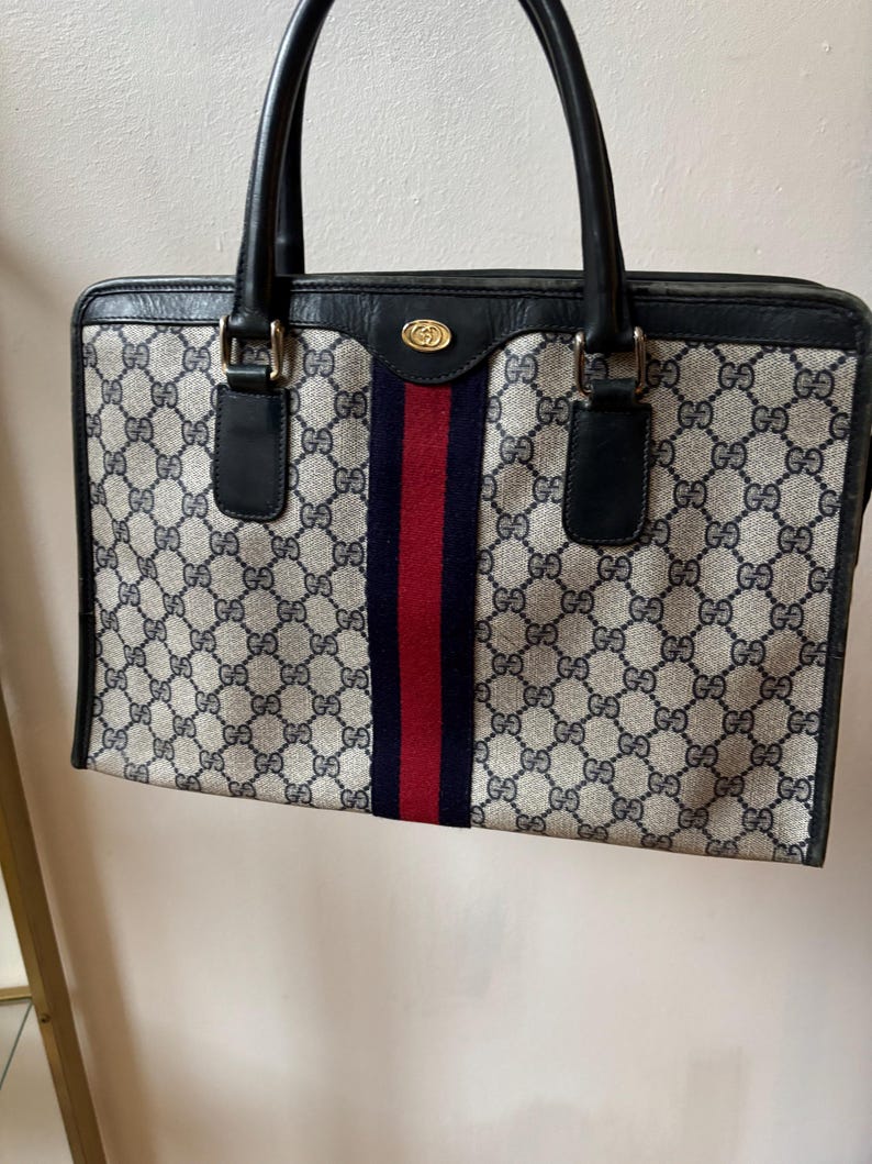 May include: A vintage handbag with a classic design. The bag features a repeating pattern of interlocking "G" logos in a beige and navy blue color scheme. A central stripe in red and navy blue adds a pop of color. The bag has black handles and trim.