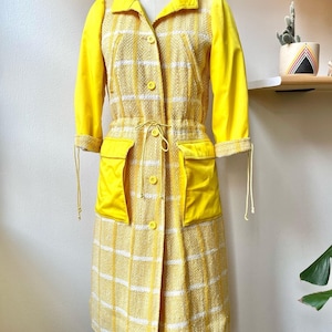 Könnte beinhalten: Ein gelbes Kleid mit weißen Karos und einer gelben Jacke mit langen Ärmeln. Das Kleid hat eine Kordeltaille und zwei große Taschen.