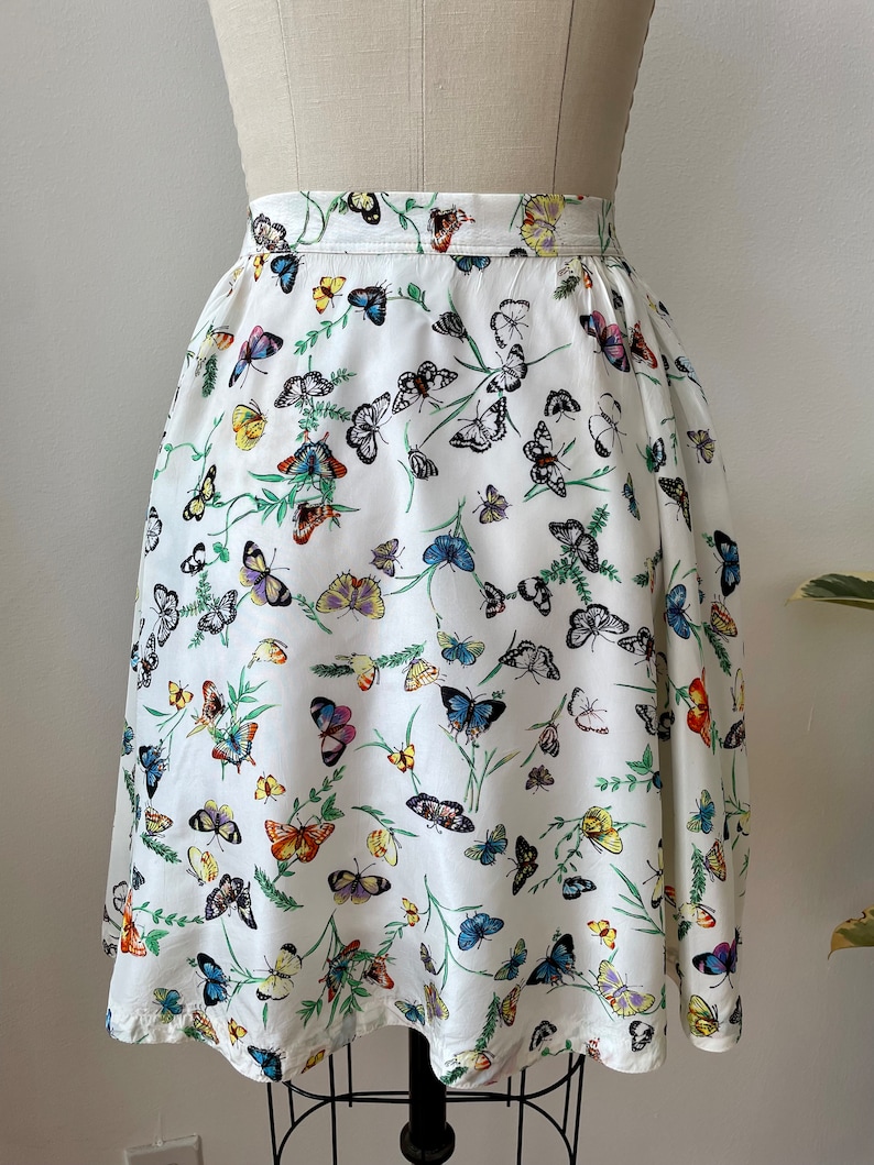 May include: A white skirt with a butterfly print. The butterflies are in various colors, including blue, yellow, orange, and green. The skirt is a-line and has a gathered waist.